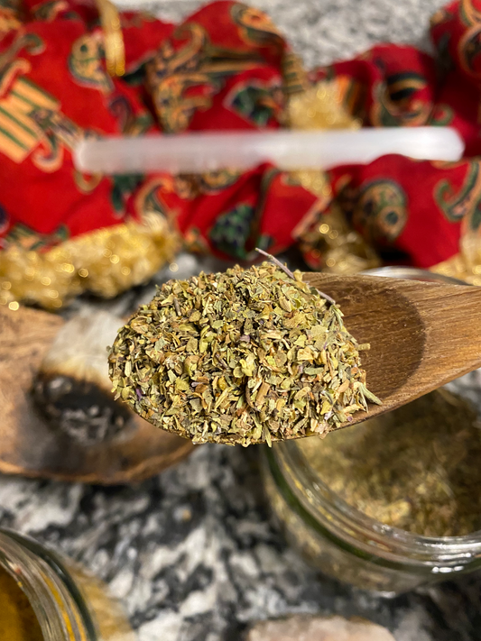 green herbal spice blend in a wooden spoon. there is a glass jar with more of the blend, and a red scarf and crystals in the picture as well.