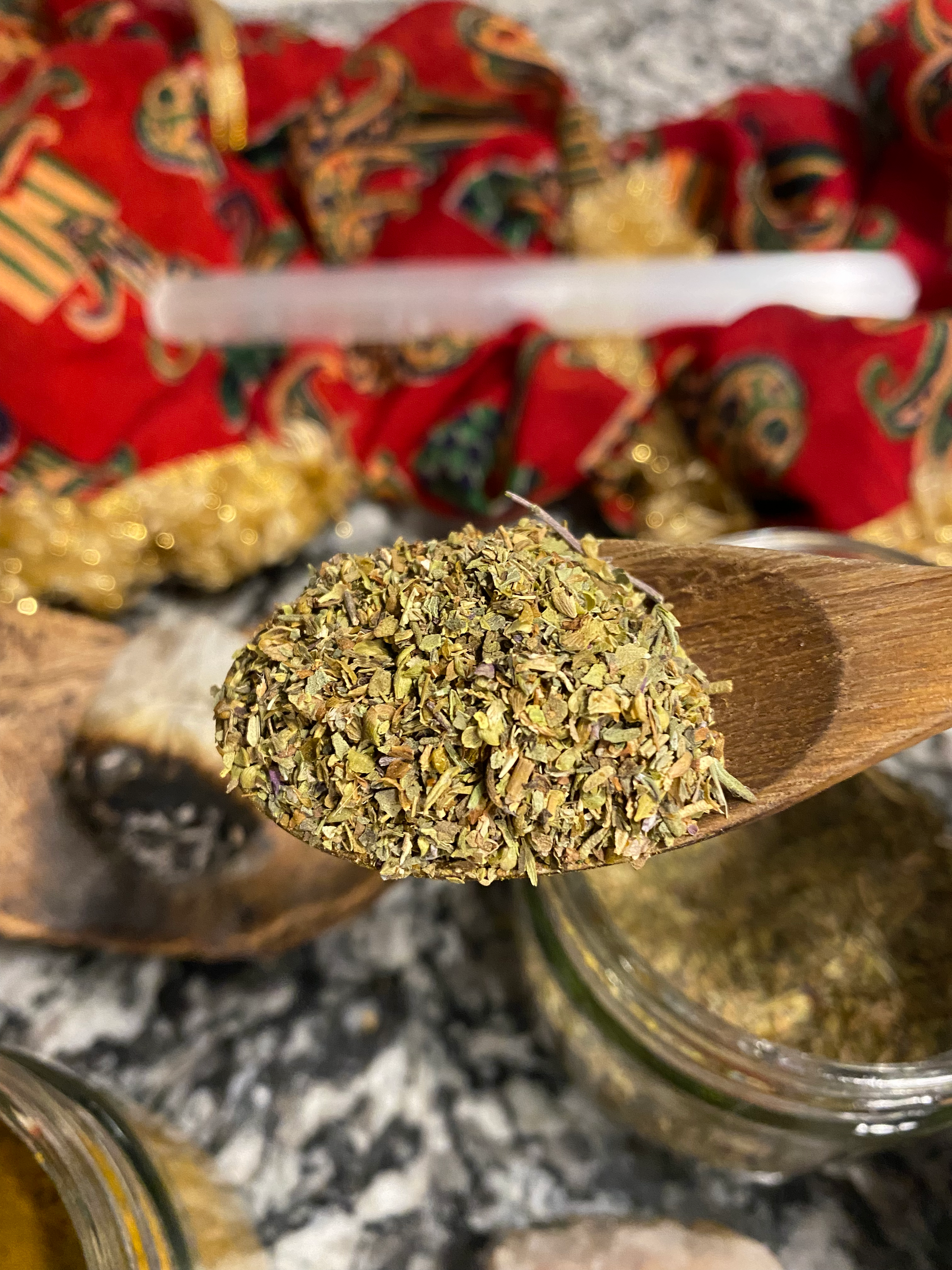 green herbal spice blend in a wooden spoon. there is a glass jar with more of the blend, and a red scarf and crystals in the picture as well. 
